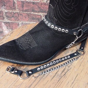 May include: Black leather cowboy boot with a silver studded strap and a chain attached to a leather strap. The boot is sitting on a wooden surface.