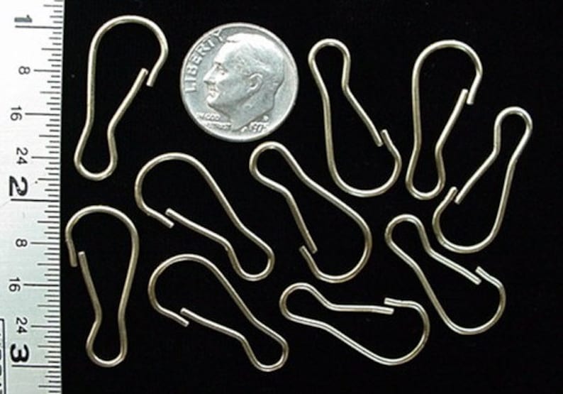 May include: Nine silver metal curtain rings, each with a rounded oval shape and a small hook at the top. The rings are arranged in a random pattern on a black surface. A US dime is included for scale.