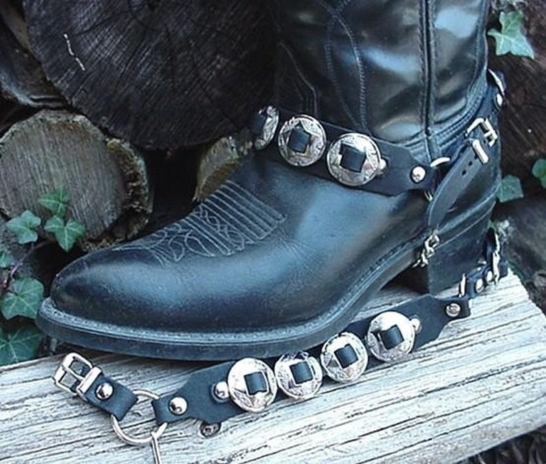 Puede incluir: Bota de cuero negro con detalles de concho plateado en la correa del tobillo y una correa de cuero negro con conchos plateados alrededor del tal&oacute;n.