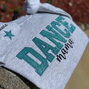 May include: A light gray hoodie with the words "DANCE mama" in teal glitter lettering. The hoodie also features a star and the name "Leena" on the hood. The hoodie is made of a soft material.