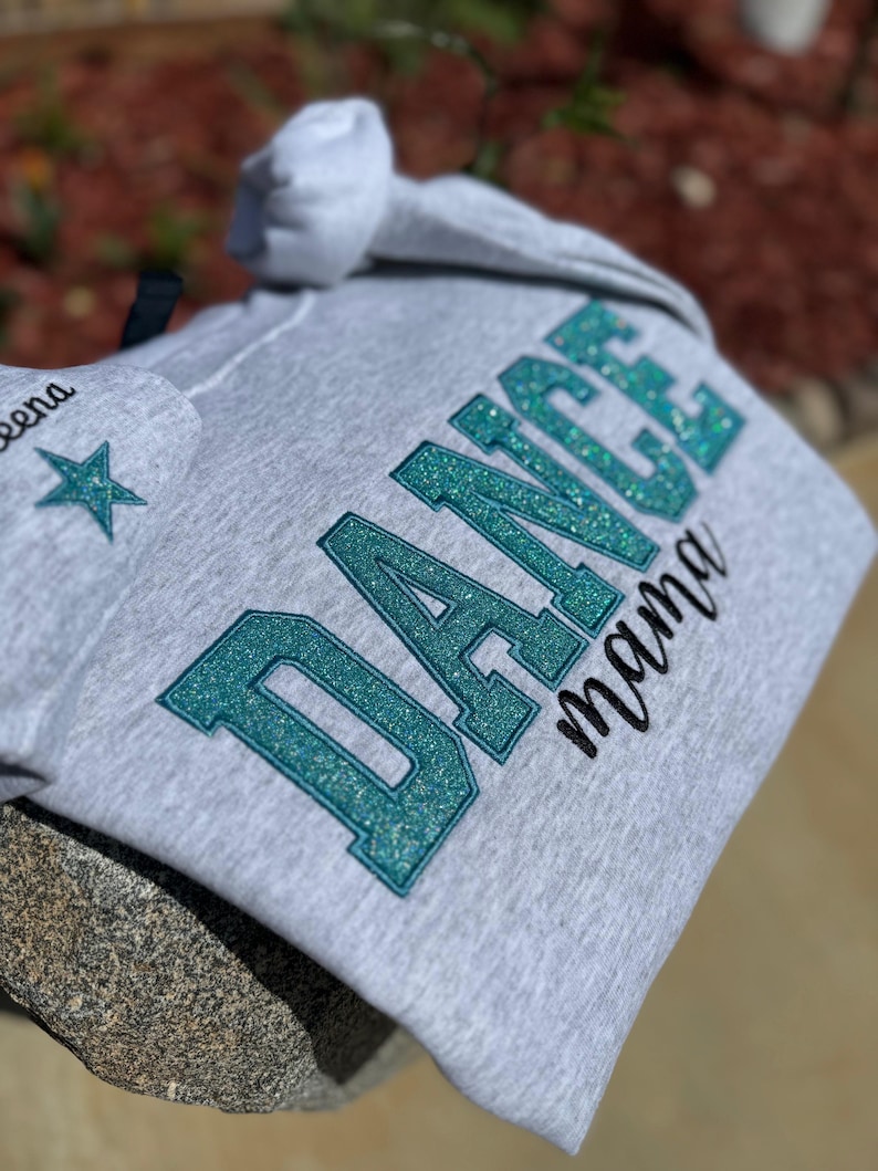 May include: A light gray hoodie with the words "DANCE mama" in teal glitter lettering. The hoodie also features a star and the name "Leena" on the hood. The hoodie is made of a soft material.