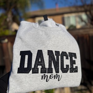May include: A light gray sweatshirt with the words "DANCE mom" in black glitter lettering. The sweatshirt is made of a soft material and is perfect for dance moms.