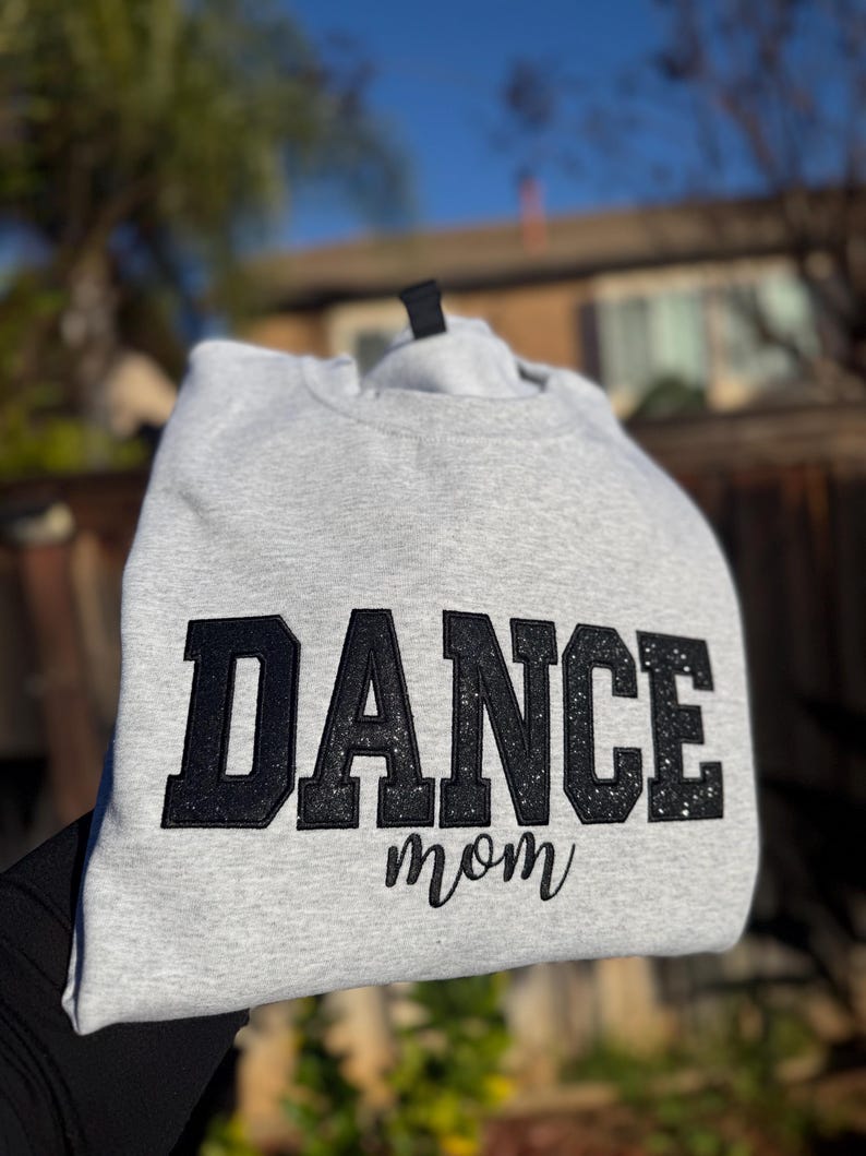 May include: A light gray sweatshirt with the words "DANCE mom" in black glitter lettering. The sweatshirt is made of a soft material and is perfect for dance moms.
