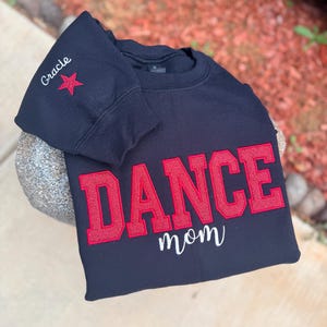 May include: A black sweatshirt with the word "DANCE" in large red letters and "mom" in white script. The sleeve has the name "Gracie" and a red star. The sweatshirt is displayed on a stone surface.