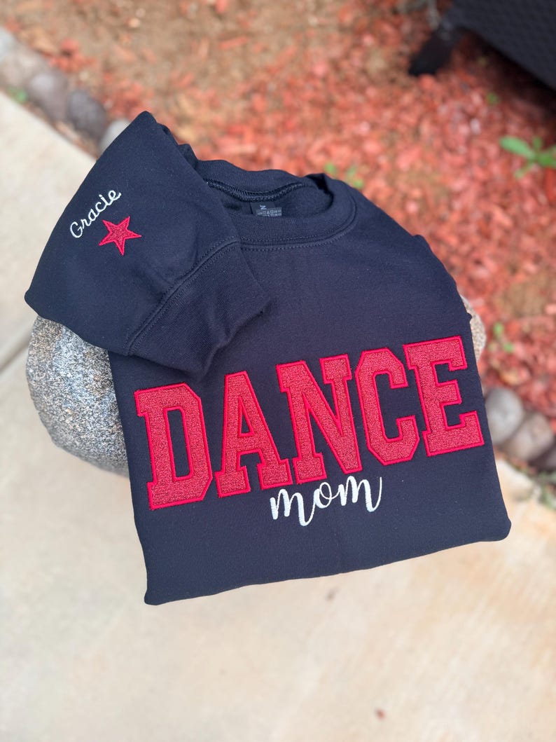 May include: A black sweatshirt with the word "DANCE" in large red letters and "mom" in white script. The sleeve has the name "Gracie" and a red star. The sweatshirt is displayed on a stone surface.