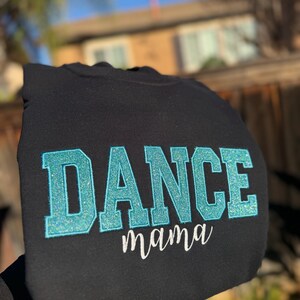 May include: Black sweatshirt with the word "DANCE" in large, glittery, turquoise letters. Below, the word "mama" is written in a delicate, white script. The sweatshirt is held up against a blurred outdoor background.