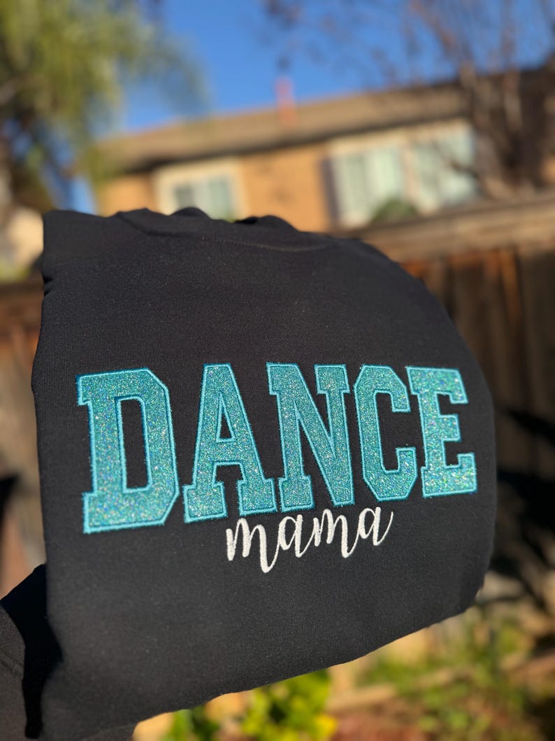 May include: Black sweatshirt with the word "DANCE" in large, glittery, turquoise letters. Below, the word "mama" is written in a delicate, white script. The sweatshirt is held up against a blurred outdoor background.