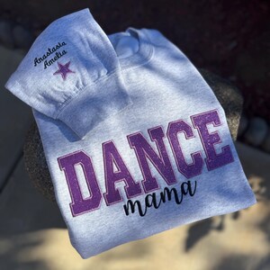 May include: A light gray sweatshirt with the words "DANCE mama" in purple glitter lettering. The sleeve has the name "Anastasia Amelia" embroidered in black with a purple star.