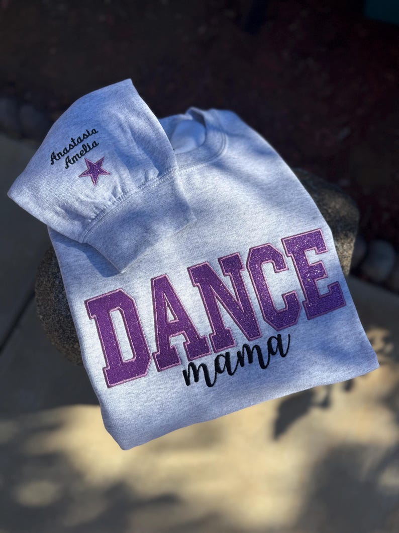 May include: A light gray sweatshirt with the words "DANCE mama" in purple glitter lettering. The sleeve has the name "Anastasia Amelia" embroidered in black with a purple star.
