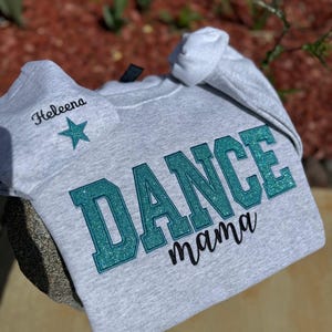 May include: A light gray sweatshirt with the words "DANCE mama" in teal glitter lettering. A matching gray beanie with the name "Heleena" and a teal star is also visible.