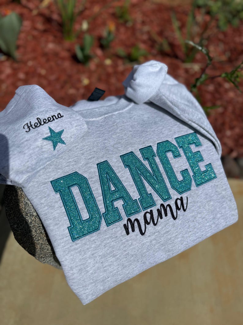 May include: A light gray sweatshirt with the words "DANCE mama" in teal glitter lettering. A matching gray beanie with the name "Heleena" and a teal star is also visible.