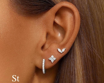 Pode incluir: Uma orelha em close-up adornada com três conjuntos de brincos. Um pequeno aro com pedras transparentes, um design de flor de quatro pétalas e um brinco em forma de marquesa. O texto "St StackedTheory" está no canto inferior esquerdo.