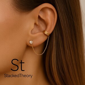 May include: Gold-toned ear cuff and stud earring set. The stud features a clear, round gemstone. A delicate chain connects the stud to the ear cuff, which has a triple-band design. The text "St StackedTheory" is visible in the image.