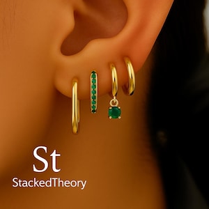 以下が含まれることがあります： 小さな緑色の宝石が付いたセットを含む、金色のフープイヤリングのコレクション。1つのイヤリングには、ぶら下がっている緑色の宝石が付いています。「St StackedTheory」の文字が画像に表示されています。