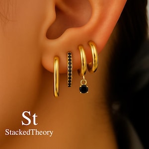 Op de afbeelding: Goudkleurige oorringen in verschillende stijlen. Eén paar is een simpele ring, een ander heeft een rij zwarte stenen en een derde heeft een hangende zwarte edelsteen. De tekst "St StackedTheory" is zichtbaar.