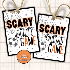 May include: Two rectangular Halloween-themed tags with the words "SCARY GOOD GAME" in black and orange. Each tag features a soccer ball and a spider web design. The tags are tied with twine.