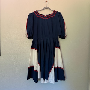 May include: A navy blue dress featuring short puff sleeves, a round neckline, and a flared skirt. The garment is accented with white panels and a red zigzag trim along the neckline, sleeves, and skirt sections, creating a distinct color-blocked look.