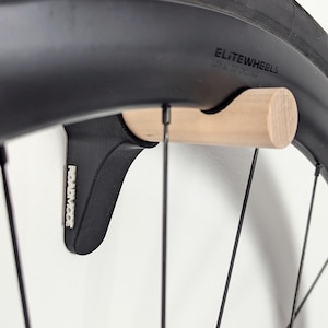 Puede incluir: Un soporte negro para rueda de bicicleta con un pasador de madera natural. El soporte está montado en una pared y sostiene un neumático negro de bicicleta. El texto "ELITEWHEELS" es visible en el neumático.