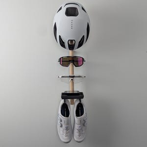 Peut inclure: Un organisateur mural pour équipements sportifs présentant un casque de vélo blanc, des lunettes de soleil et des chaussures de cyclisme. Le casque a des détails noirs et les lunettes ont des verres roses. Les chaussures sont blanches avec une fermeture à molette.