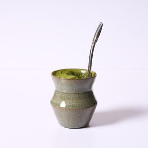 May include: A ceramic yerba mate gourd with a green interior filled with green powder and a metal bombilla straw. The gourd has a grey and green colour scheme with a rounded shape.