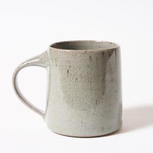 May include: A ceramic mug with a pale sage green glaze featuring a distinct crackle finish. The mug has a cylindrical body and a curved handle extending from the side, with subtle dark speckles visible throughout the glossy, textured surface.