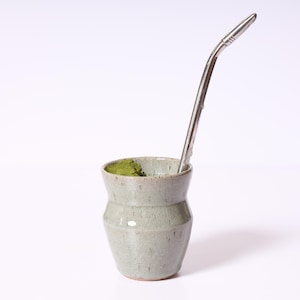 May include: A light gray ceramic mate gourd with a subtle crackle glaze finish and a tapered waist. The vessel is filled with vibrant green loose-leaf tea and features a long, slender, polished silver-toned metal bombilla straw inserted inside.