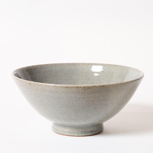 May include: A pale grey ceramic bowl with a pedestal base. The bowl has a wide, open shape and a smooth, slightly glossy surface. The rim is a darker shade of grey. Suitable for serving soups, salads, or other dishes.