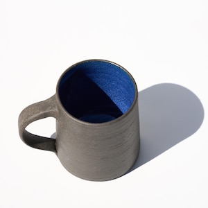 May include: A handcrafted ceramic mug with a dark grey exterior and a vibrant blue interior. The mug has a rounded shape with a sturdy handle, ideal for enjoying coffee or tea. The mug's dimensions are approximately 10 cm in height and 8 cm in diameter.