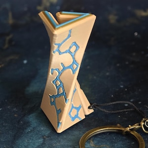 May include: A beige and blue geometric pendant with a bronze-coloured key ring. The pendant has a twisted design with blue accents. The key ring is round and has a clasp. The background is a dark blue with gold flecks.