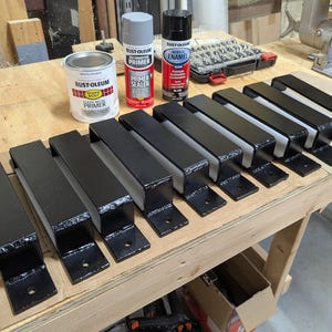May include: A collection of black metal brackets with a rectangular design, arranged on a wooden surface. Several cans of primer and enamel paint are also visible, including a can of white primer and a can of gray primer sealer.