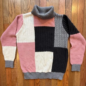 May include: A patchwork sweater with a gray turtleneck and cuffs. The sweater features blocks of pink, white, black, and gray knit fabric. The sweater is made of a soft, comfortable material.