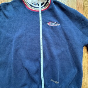 May include: Navy blue sweatshirt with the text "Field of Dreams" embroidered on the chest and lower right. The collar is white with red and black stripes. The sweatshirt is measured with a tape measure.