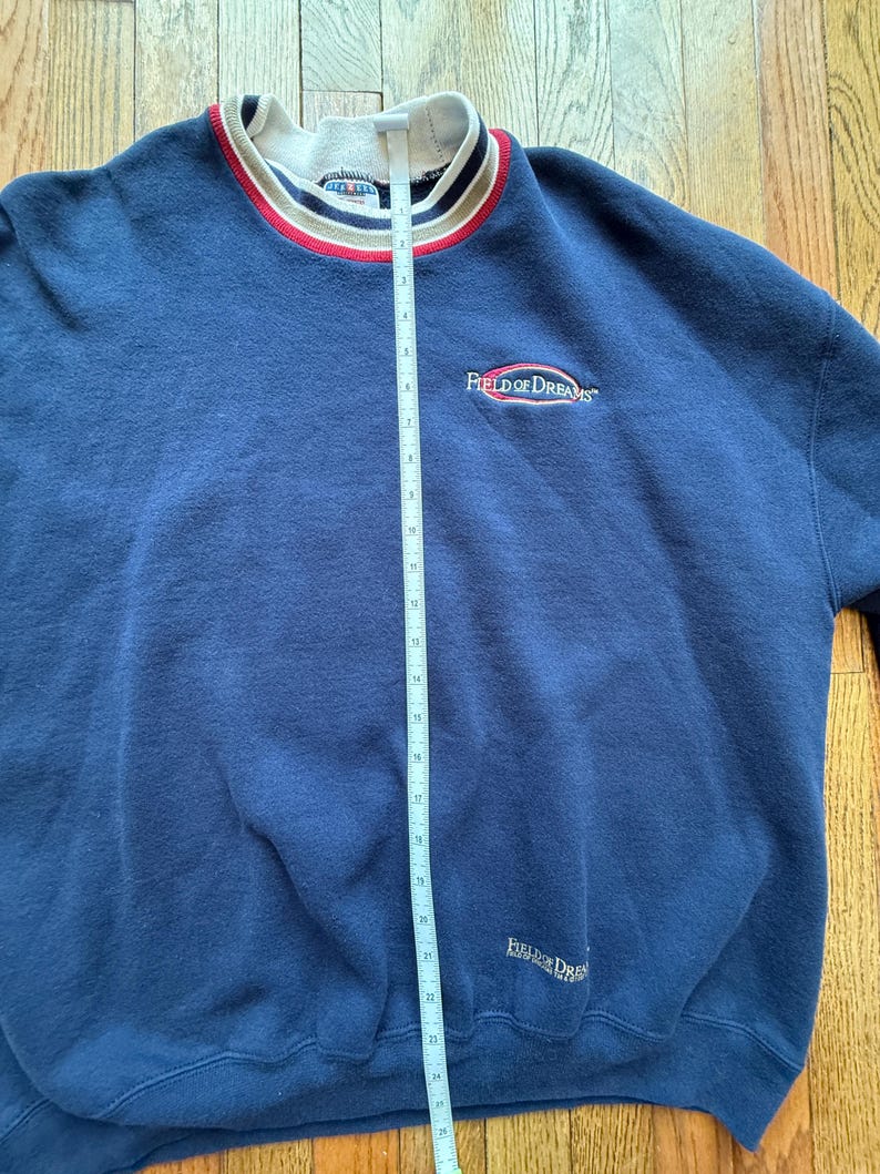 May include: Navy blue sweatshirt with the text "Field of Dreams" embroidered on the chest and lower right. The collar is white with red and black stripes. The sweatshirt is measured with a tape measure.