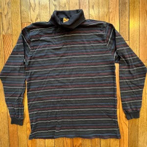 May include: A long-sleeved turtleneck shirt with horizontal stripes in red, white, blue, and green on a dark gray background. The shirt has a ribbed collar and cuffs.