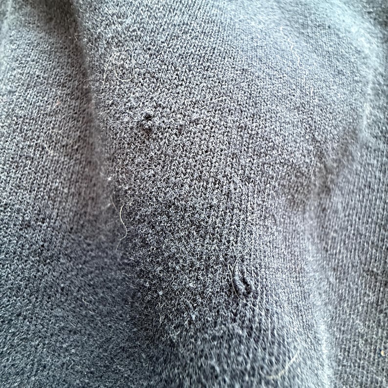 May include: Close-up of a dark gray knit fabric with visible texture and small imperfections. The fabric appears to be a soft material, possibly a sweater or other garment. The image highlights the detailed weave and subtle variations in color.