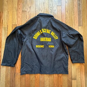May include: Black jacket with yellow text on the back. The text reads "BOONE & SCENIC VALLEY RAILROAD" above "BOONE IOWA". The jacket has a collared neckline and long sleeves.