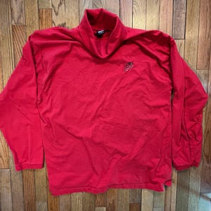May include: A bright red long-sleeved shirt with a mock turtleneck collar. The shirt has a small embroidered design on the upper left chest. The shirt is made of a soft, comfortable material.