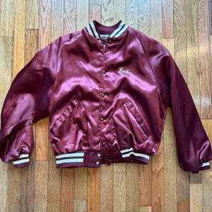 May include: A maroon satin bomber jacket with white and maroon striped trim on the collar, cuffs, and hem. The jacket has a button-up front and two front pockets. The name "Bert" is embroidered on the chest.