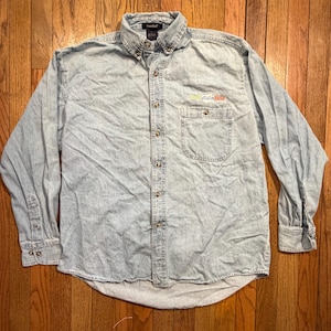 May include: A light blue denim long-sleeve button-down shirt with a collar and a single chest pocket. The shirt features a small embroidered logo above the pocket that reads "Iowa State Fair" in multi-colored lettering. It has metal buttons.