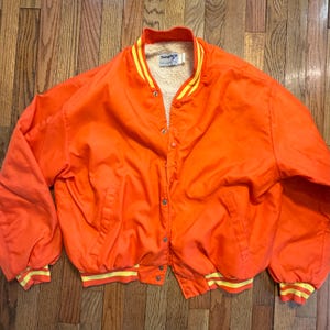 May include: A bright orange varsity jacket featuring a cream-colored sherpa lining, a snap-button front closure, and ribbed cuffs and collar with yellow stripes. The interior label displays the brand name Swingster in blue text.