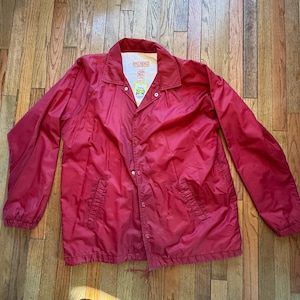 May include: A red coach's jacket with a white collar and snap closures. The jacket has two front pockets and a drawstring at the bottom. The Big Mac logo is visible on the inside collar.