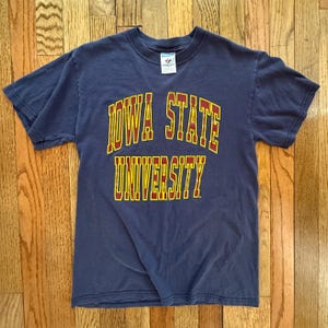 May include: Navy blue short-sleeved t-shirt with the words "IOWA STATE UNIVERSITY" in gold and red lettering. The shirt is made of a soft material and is likely vintage.