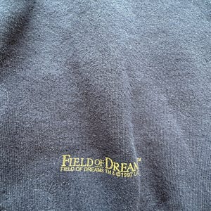 May include: Navy blue sweatshirt with the text "FIELD OF DREAMS" in gold lettering. The text "FIELD OF DREAMS TM & &copy;1997 U" is printed below the main text. The sweatshirt has a textured appearance.