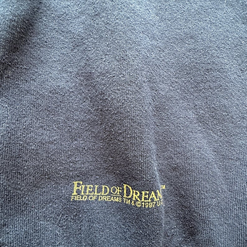 May include: Navy blue sweatshirt with the text "FIELD OF DREAMS" in gold lettering. The text "FIELD OF DREAMS TM & &copy;1997 U" is printed below the main text. The sweatshirt has a textured appearance.