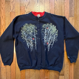 May include: A navy blue button-up sweatshirt with a red collar. The sweatshirt features a design of white trees with yellow leaves and red birds. The sweatshirt has two pockets and long sleeves.