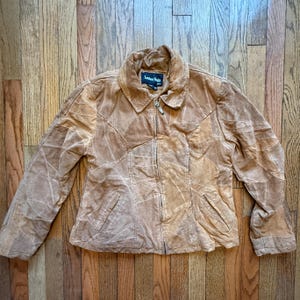 May include: A tan suede jacket with a patchwork design. The jacket has a collared neckline, a zipper closure, and two front pockets. The label reads "Leather Works".