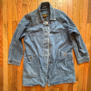 May include: A light blue denim jacket with a collared neckline, button closures, and two front pockets. The jacket has long sleeves and a classic, casual style. The jacket is laid flat on a wooden surface.
