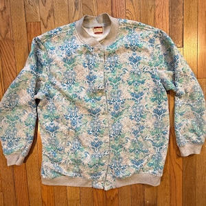 May include: A button-up bomber jacket with a gray collar, cuffs, and waistband. The jacket features a repeating pattern in shades of blue and teal on a gray background. The jacket has two front pockets.