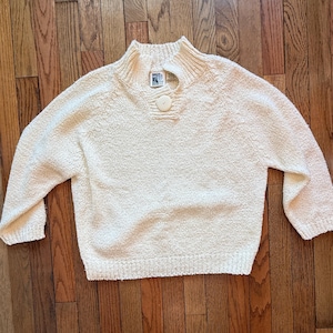 May include: Cream-colored sweater with a high collar and a button closure at the neckline. The sweater has long sleeves and a textured knit design. The garment is made of a soft, warm material, perfect for cooler weather.