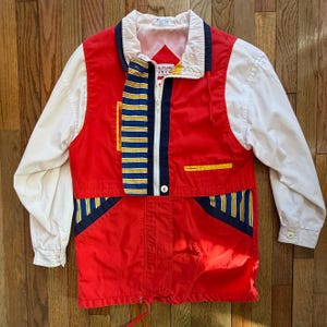 May include: A vintage red, white, and blue jacket with a zip-up front. The jacket features white sleeves, a white collar, and striped accents in blue and yellow. The jacket has a drawstring at the bottom.